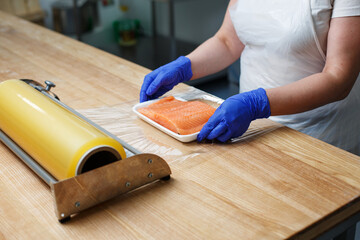Worker in factory or shop packs raw fresh salmon fillets in cling film.
