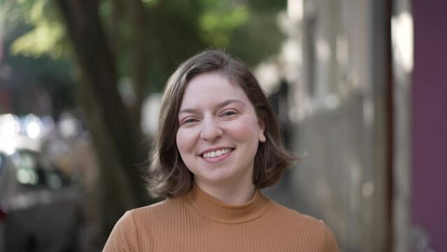 Portrait of a joyful woman walking toward camera. Tracking motion shot of person coming forward smiling. Happy millennial 20s girl in city sidewalk
