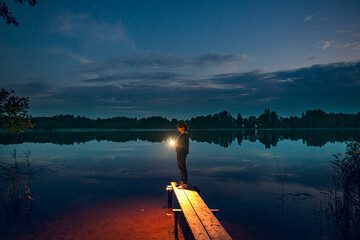 a young girl with a flashlight stands on the shore of a forest lake, as the lighthouse illuminates the way to the shore. Waiting for a guy from fishing. evening comes,the first stars appear in the sky - obrazy, fototapety, plakaty
