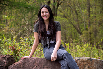 Casual lifestyle outdoor portrait of a beautiful happy young woman with long brown hair sitting on rocks smiling 