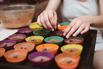 A young teenage girl makes homemade cupcakes at home, pours the dough into special rubber molds and puts chocolate, close up, focus on hands