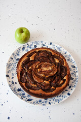 A nordic style apple tart on a blue and white plate against a white speckled background. A single apple i seen in the top left corner.