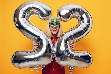 a happy emotional woman stands in a red shirt with green festive glasses, holding balloons in the form of the number twenty-two on a yellow background, her mouth wide open with delight