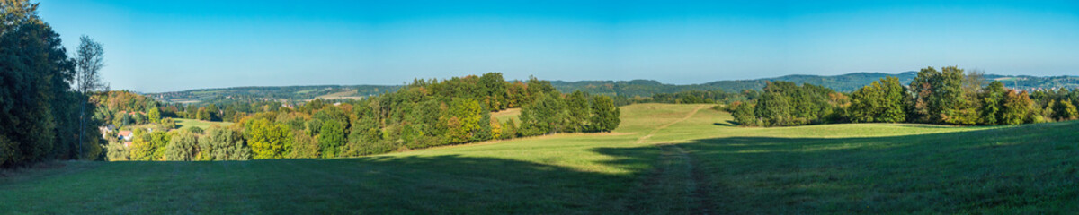 Ultra wide panoramic late summer landscape with idyllic green meadow, trees, forest and hills with...