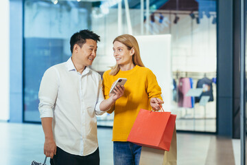 Diverse couple Asian man and blonde woman in clothes supermarket, looking at mobile phone screen, choosing shopping, holding colorful shopping bags and gifts.