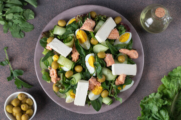 Salad with canned salmon, egg, cucumber, arugula and feta cheese on round plate on brown background