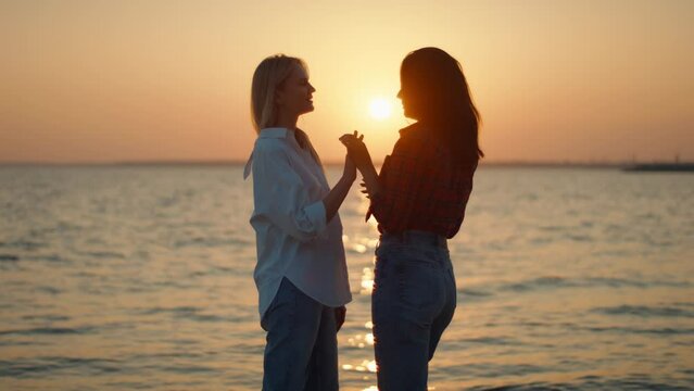 Silhouette of homosexual lesbian couple of two young women by sea against backdrop of sunset. Gentle hugs and touches of couple in love
