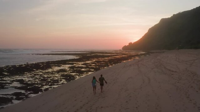 A couple walking along sandy beach at sunset. Adult man and woman strolling on a seaside with white sand and reef towards hills at dusk. Aerial drone camera footage of holiday-makers at tropical shore