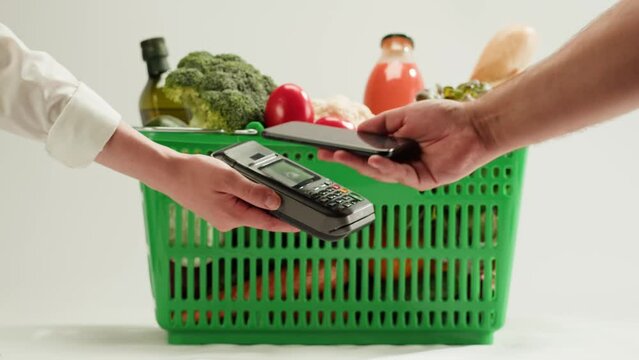 Food shopping delivery, paying with smartphone in shop. Buying eats in supermarket store. Green basket with fresh vegetables, goods products. 