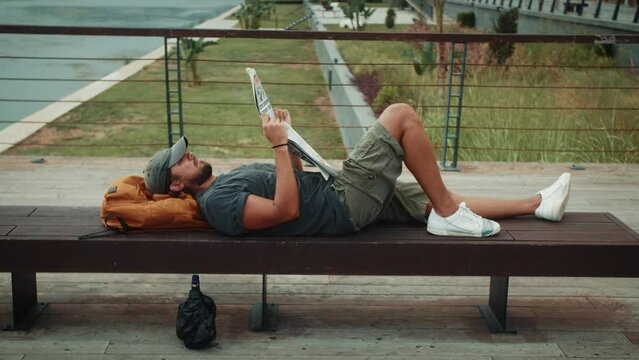 Young tourist man lying on bench in park reading a newspaper on a sunny day. Homeless sleep on backpack