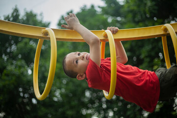 Boy on playground climbs through rings. Child plays at height. Sports ground for children to play. Boy shows sports skills. © Олег Копьёв