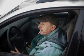 A man with a cigarette driving a car. The guy in the cap in his car.