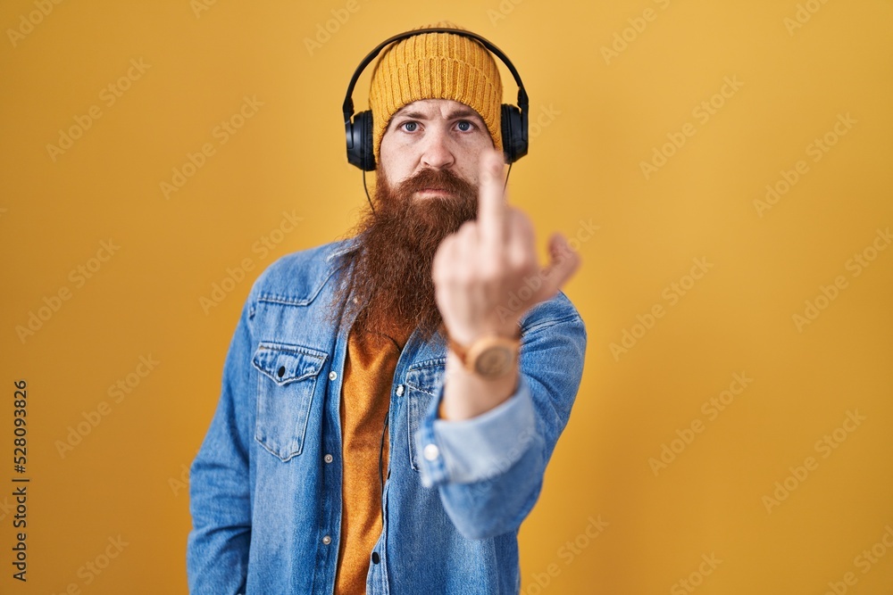 Wall mural caucasian man with long beard listening to music using headphones showing middle finger, impolite an