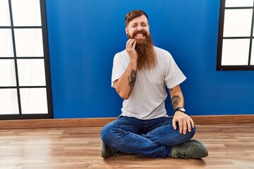 Redhead man with long beard sitting on the floor at empty room touching mouth with hand with painful expression because of toothache or dental illness on teeth. dentist