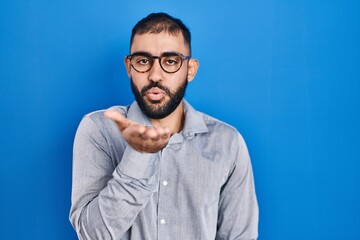 Middle east man with beard standing over blue background looking at the camera blowing a kiss with hand on air being lovely and sexy. love expression.