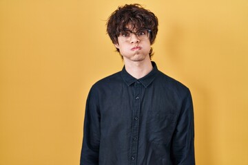 Obraz premium Young man wearing glasses over yellow background puffing cheeks with funny face. mouth inflated with air, crazy expression.