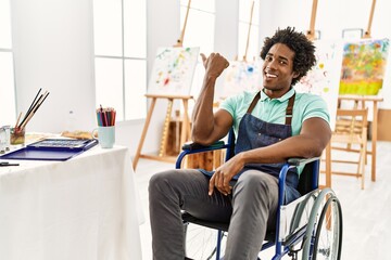 Young african american artist man sitting on wheelchair at art studio smiling with happy face looking and pointing to the side with thumb up.