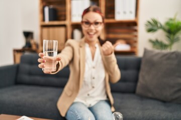 Young caucasian woman offering a glass of water pointing thumb up to the side smiling happy with open mouth