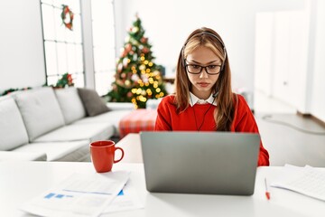 Young caucasian girl sitting on the table working using laptop by christmas tree relaxed with serious expression on face. simple and natural looking at the camera.