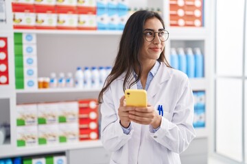 Young hispanic girl pharmacist using smartphone working at pharmacy