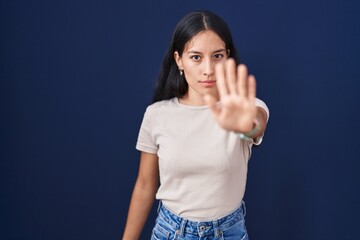 Fototapeta premium Young hispanic woman standing over blue background doing stop sing with palm of the hand. warning expression with negative and serious gesture on the face.