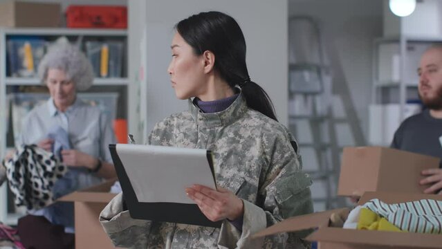 Refugees support. Female military officer with clipboard supervise work of volunteers in warehouse. Diverse people sort and pack donated goods for humanitarian aid