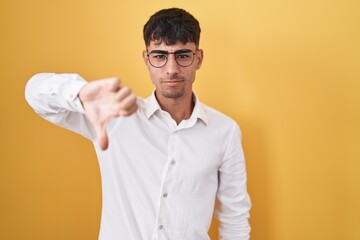 Young hispanic man standing over yellow background looking unhappy and angry showing rejection and negative with thumbs down gesture. bad expression.
