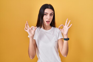 Young beautiful woman standing over yellow background looking surprised and shocked doing ok approval symbol with fingers. crazy expression