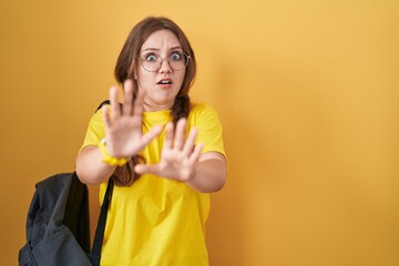 Young caucasian woman wearing student backpack over yellow background afraid and terrified with fear expression stop gesture with hands, shouting in shock. panic concept.