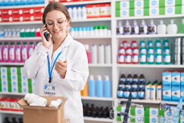 Young blonde woman pharmacist holding pills bottle talking on smartphone at pharmacy