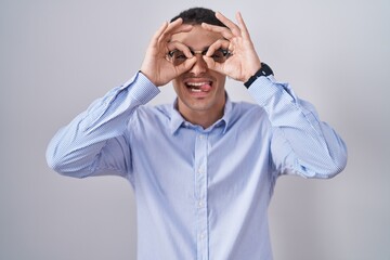Handsome hispanic man wearing business clothes and glasses doing ok gesture like binoculars sticking tongue out, eyes looking through fingers. crazy expression.