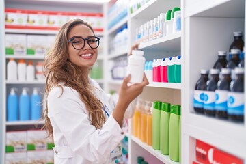 Young beautiful hispanic woman pharmacist smiling confident holding pills bottle on shelving at pharmacy