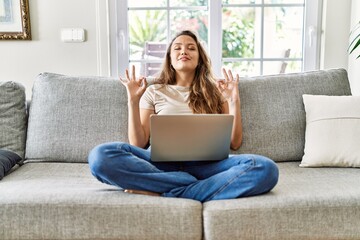Beautiful young brunette woman sitting on the sofa using computer laptop at home relax and smiling with eyes closed doing meditation gesture with fingers. yoga concept.