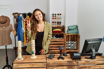 Beautiful hispanic woman working at fashion shop showing and pointing up with finger number one while smiling confident and happy.