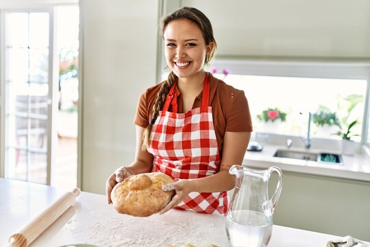 Young beautiful hispanic woman smiling confident holding bread at the kitchen