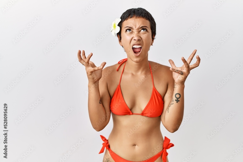 Sticker Young hispanic woman with short hair wearing bikini crazy and mad shouting and yelling with aggressive expression and arms raised. frustration concept.