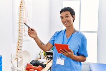 Young hispanic woman wearing physiotherapist uniform pointing to vertebral column using touchpad at...
