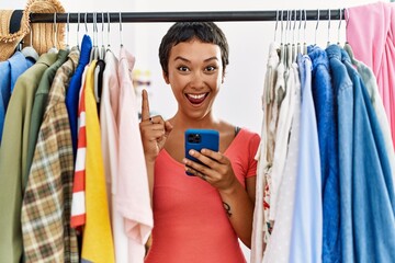 Naklejka na ściany i meble Young hispanic woman with short hair searching clothes on clothing rack using smartphone surprised with an idea or question pointing finger with happy face, number one