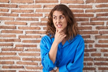 Beautiful brunette woman standing over bricks wall thinking worried about a question, concerned and nervous with hand on chin
