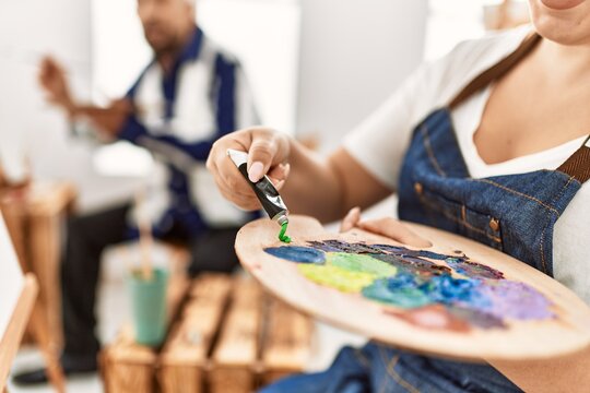 Young artist woman and senior painter man at art studio classroom painting on canvas, close up of woman pouring oil paint on artist palette