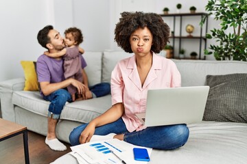 Mother of interracial family working using computer laptop at home puffing cheeks with funny face. mouth inflated with air, crazy expression.