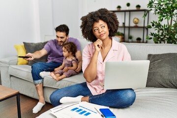 Mother of interracial family working using computer laptop at home with hand on chin thinking about question, pensive expression. smiling with thoughtful face. doubt concept.
