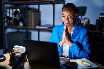 Beautiful african american woman working at the office at night praying with hands together asking...