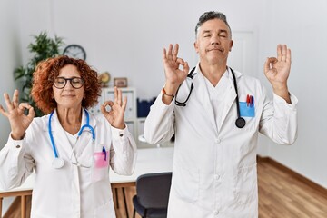 Two middle age doctors at medical clinic relax and smiling with eyes closed doing meditation gesture with fingers. yoga concept.