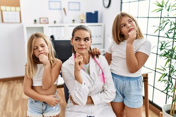 Pediatrician woman working at the clinic with two little girls serious face thinking about question...