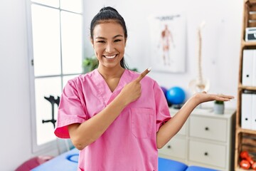Fototapeta premium Young hispanic woman working at pain recovery clinic amazed and smiling to the camera while presenting with hand and pointing with finger.