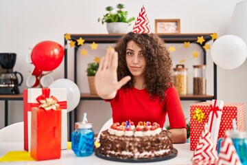 Hispanic woman with curly hair celebrating birthday holding big chocolate cake with open hand doing stop sign with serious and confident expression, defense gesture