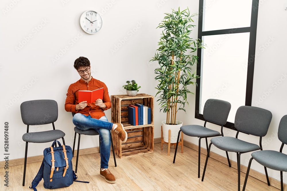 Sticker young hispanic man smiling confident reading book at waiting room