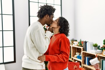 Young interracial couple kissing and hugging standing by christmas tree at home.