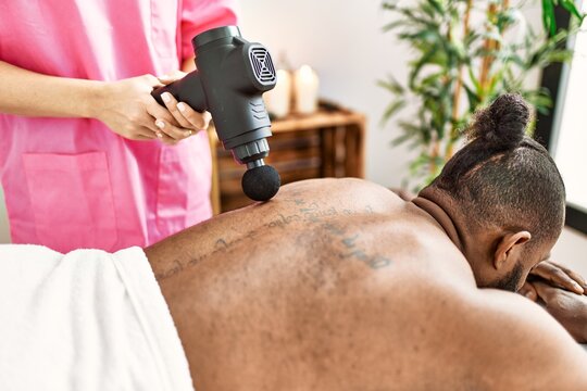 Young african american man having back massage with percussion pistol at beauty center